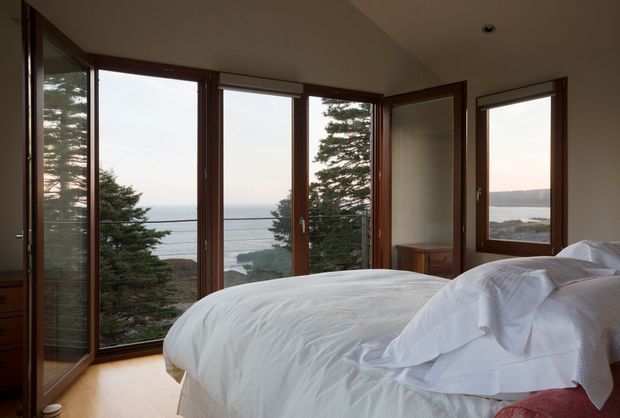 A serene bedroom with a large bed positioned beside expansive timber-framed corner glazing and opening glass doors. The windows overlook trees, water, and a distant shoreline, bringing abundant natural scenery into the room. The interior has warm timber flooring and window frames, pale walls, simple white bedding, and a restrained contemporary design.