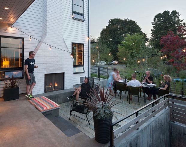 An elevated outdoor dining and entertaining terrace beside a contemporary white-painted brick and weatherboard-clad building. The space includes a built-in outdoor fireplace, dining table with guests, lounge seating, potted plants, festoon lighting, dark metal railings, and paved flooring overlooking a landscaped suburban setting.