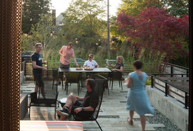 A landscaped outdoor terrace or rooftop-style deck with several people gathered around a dining table. The space features timber decking, black metal railings and furniture, festoon lighting, concrete edging, lush planting, and a prominent red-leaved ornamental tree. A patterned vertical screen frames the left side, creating a relaxed social outdoor setting.