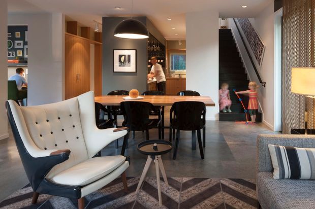 A warm, contemporary open-plan living and dining area with a timber dining table, black dining chairs, sculptural pendant light, and a prominent white-and-black lounge chair. The space opens toward a kitchen and hallway, with a staircase at right and patterned flooring creating a strong geometric feature. Built-in timber joinery, dark cabinetry, and a perforated metal or mesh screen add texture.
