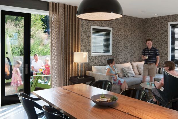 A bright, open-plan dining and living area with a large solid timber dining table and black chairs in the foreground. A sectional sofa, floor lamp, patterned wallpaper, horizontal blinds, and a large dark pendant light define the living zone. Full-height glazed doors open to an outdoor area, with family members visible inside and outside.