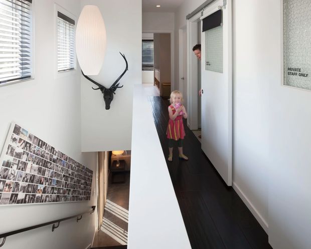 Bright narrow interior hallway with white walls, dark timber flooring, multiple windows fitted with white horizontal blinds, and white doors. A long photo collage runs along the stairwell wall beside a dark handrail, while a sculptural black antler wall mount and elongated white pendant add decorative focal points. A sign identifies a private staff-only area, suggesting a workplace or commercial interior.