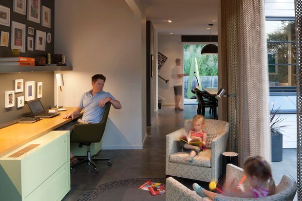 A warm, contemporary open-plan home interior showing a compact built-in home office along a hallway, with a desk, computer, shelving and framed artwork. The corridor opens toward a dining area and garden-facing glazing, while two children sit in patterned armchairs near the window. Dark flooring, pale cabinetry, neutral walls, and layered indoor lighting create a relaxed family setting.