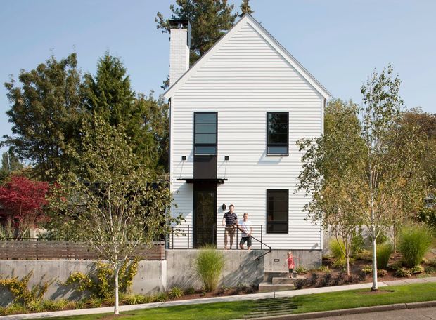 Front exterior view of a narrow, multi-storey house with a steep front-facing gable roof, white horizontal weatherboard cladding, dark-framed windows, and a tall white chimney. The home is set behind a landscaped frontage with young trees, grasses, retaining walls, a concrete entry stair, and a pedestrian sidewalk. The simple gabled form and compact urban frontage are the dominant architectural features.