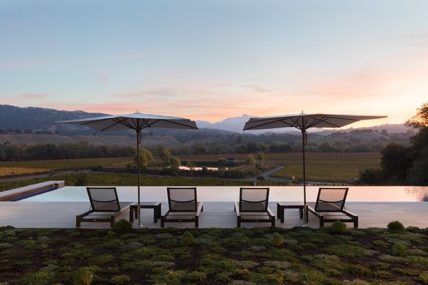 A tranquil outdoor terrace overlooking cultivated fields and a mountain range at sunset. A long reflective pool or water feature runs along the terrace edge, furnished with four lounge chairs, small side tables, and two large cantilevered umbrellas. The scene has a low-profile, refined resort-like design integrated into a rural landscape.