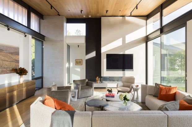 A bright contemporary living room with a large sectional sofa, additional lounge chairs, a round coffee table, and a wall-mounted television above a linear fireplace or media console. The room features floor-to-ceiling glazing, strong natural light, a warm timber ceiling, dark window frames, pale textured walls, and an open double-height volume.