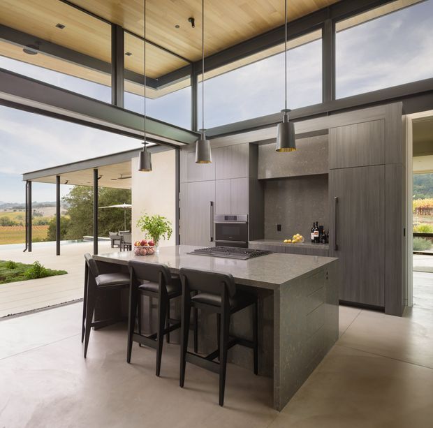 A contemporary open-plan kitchen with a large stone island and seating for four, dark integrated cabinetry, a textured stone backsplash, and a warm timber-lined ceiling. Full-height glazing and wide openings connect the kitchen to a covered outdoor terrace and landscaped setting. Black-framed glazing, exposed structural beams, pendant lights, and polished concrete flooring create a refined indoor-outdoor composition.