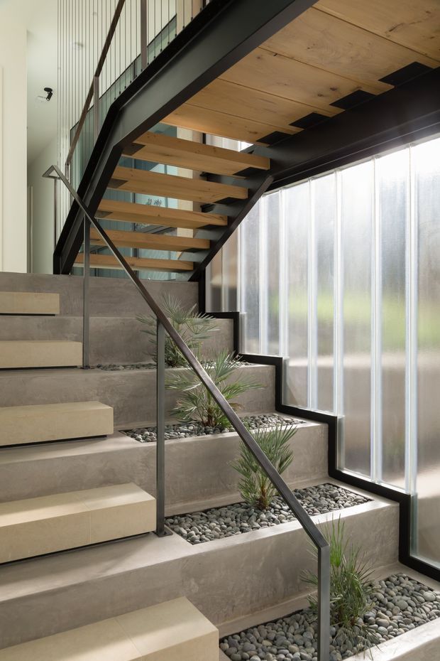 A contemporary interior stairway with floating timber treads and dark metal stringers and handrail. The stair rises through a tall, light-filled space beside translucent floor-to-ceiling glazing, with integrated concrete planter beds filled with smooth river stones and small palms. A vertical slatted wall and upper-level balustrade are visible in the background.