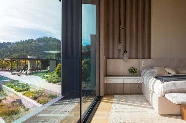 Contemporary bedroom opening onto a landscaped pool terrace through full-height sliding glass doors. The room features a low upholstered bed, pale bedding, a built-in bedside cabinet, a vertically ribbed timber feature wall, hanging pendant lights, and warm light timber flooring. The glazing provides views of the outdoor pool area and wooded hillside beyond.