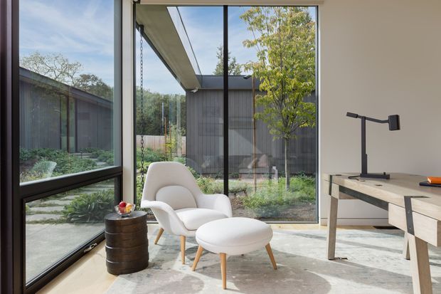 A bright, minimalist sitting area beside full-height glazing, furnished with a white upholstered lounge chair and ottoman, a small dark round side table, and a simple timber desk. The room opens visually to a landscaped courtyard and a dark vertically clad pavilion-like structure outside. Black-framed windows, pale walls, a light concrete or stone floor, and clean-lined furniture create a calm contemporary interior.
