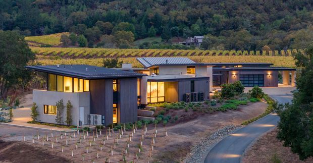A contemporary single-storey residential compound set within a rural vineyard landscape. Several low-profile buildings are arranged around landscaped planting and paved access paths, with dark metal-clad forms, pale rendered walls, broad glazed openings, and flat or very low-pitched roofs. The buildings are photographed at dusk with warm interior and exterior lighting, while rows of young vines occupy the foreground and mature wooded hills rise behind.
