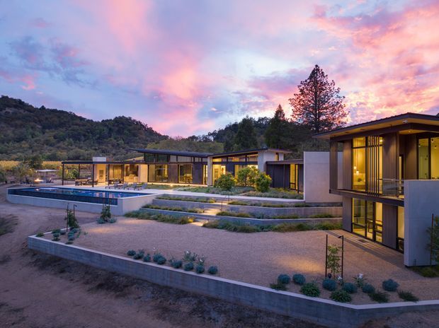 A contemporary multi-wing residence set into a landscaped hillside at dusk. The low-profile buildings feature flat roofs, extensive floor-to-ceiling glazing, dark window frames, warm interior lighting, and pale rendered or concrete walls. Terraced gravel gardens with planted borders lead to a rectangular swimming pool, while wooded hills rise behind the property beneath a vivid pink and blue sunset sky.
