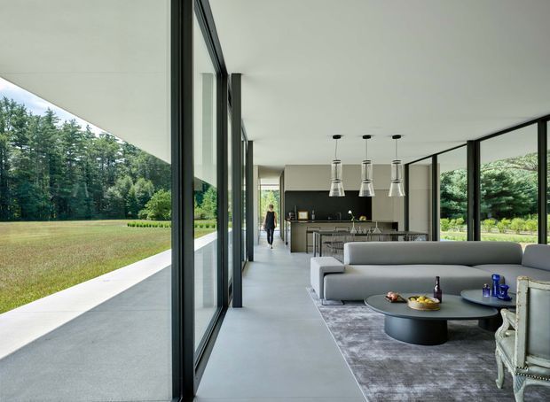 A spacious, contemporary open-plan living area with floor-to-ceiling black-framed glazing opening onto a landscaped lawn and tree-lined grounds. The room includes a grey sectional sofa, dark rounded coffee tables, a large area rug, pendant lights, and a dining area leading toward a sleek kitchen with dark cabinetry and a central island. The long, low form, expansive glazing, and restrained palette create a calm indoor-outdoor connection.