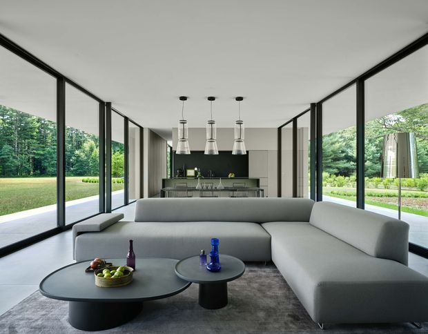 A spacious, minimalist open-plan living room with floor-to-ceiling black-framed glazing on three sides, overlooking a manicured green landscape. A large light-grey sectional sofa faces two round black coffee tables, while a compact modern kitchen with a dark backsplash and pendant lights sits beyond the seating area. The room has a white ceiling, pale flooring, restrained furnishings, and strong indoor-outdoor visual connection.