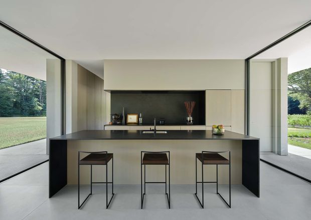 A minimalist contemporary kitchen viewed straight on, with a large island and three slim bar stools centered in the room. The kitchen features pale integrated cabinetry, a dark stone-look benchtop and matching dark side panels, a dark splashback, and extensive floor-to-ceiling glazing on both sides opening toward a landscaped exterior. The space has a flat, clean-lined ceiling and a restrained neutral palette.