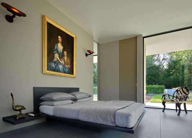 A contemporary bedroom with a low-profile dark upholstered platform bed, layered grey bedding, and large-format pale floor tiles. Full-height glazing and an open glass door connect the room to a leafy outdoor setting. A traditional gold-framed portrait and sculptural wall lights add contrast to the otherwise minimal interior.