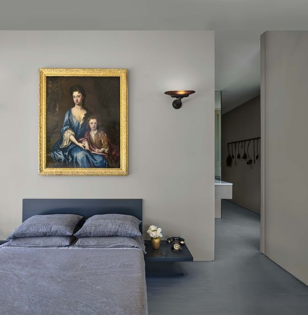 A refined bedroom with a low-profile dark upholstered bed and muted blue-grey bedding. The room has smooth light-grey walls, dark grey flooring, a compact floating bedside table, and a traditional gold-framed portrait above the bed. A wall sconce provides warm accent lighting, while an open passage reveals a minimalist adjoining circulation area with wall-mounted hooks and partial built-in cabinetry.