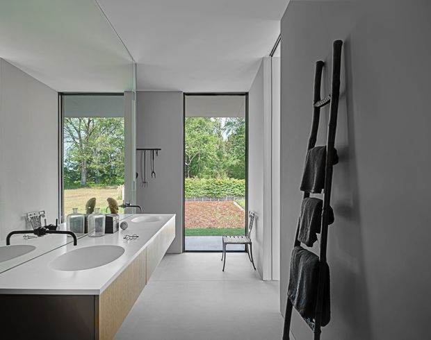 Contemporary minimalist bathroom with a long white double vanity, integrated oval basins, pale wood cabinetry, large frameless or slim-framed windows, and a glass door opening to a landscaped garden. A dark leaning towel ladder provides contrast against the light grey and white interior.
