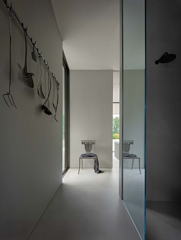 A narrow, minimalist interior corridor leading toward a bright sitting area, with smooth grey walls and pale flooring. A sculptural metal chair is positioned at the end of the passage and reflected in a full-height glass or mirrored partition on the right. The left wall features a row of dark decorative hooks holding sculptural objects, while a black shower fixture is visible through the glass on the right, suggesting an adjacent bathroom or wet area.