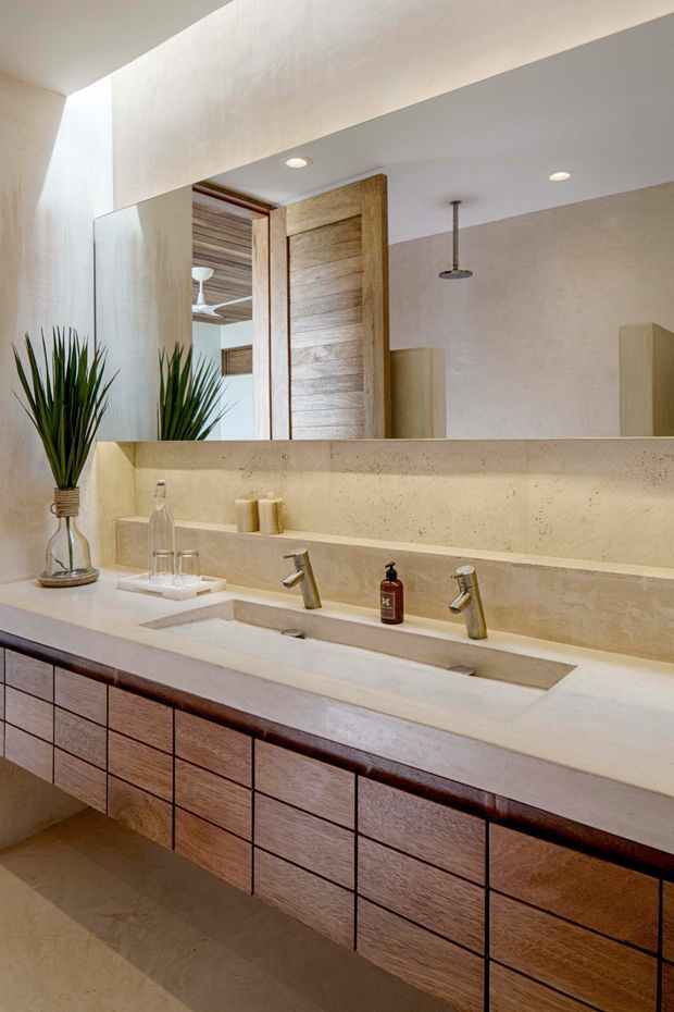 A warm, refined bathroom vanity with a long double basin set into a pale stone or concrete benchtop. The wall behind the vanity has a matching integrated ledge and large mirror, while dark timber drawer cabinetry provides contrast below. A timber pivot door, recessed ceiling lights, and a ceiling-mounted shower are visible in the reflection and adjoining area. Natural greenery and restrained accessories add an organic touch.