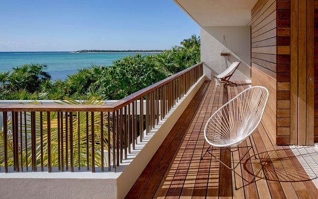 A sunny elevated balcony or terrace overlooking vivid turquoise ocean water and lush tropical vegetation. The space features warm timber decking and horizontal timber wall cladding, a dark timber-and-metal balustrade, white rendered walls, and lightweight wire lounge chairs. The long covered balcony has an airy resort-like coastal atmosphere with strong indoor-outdoor connection.