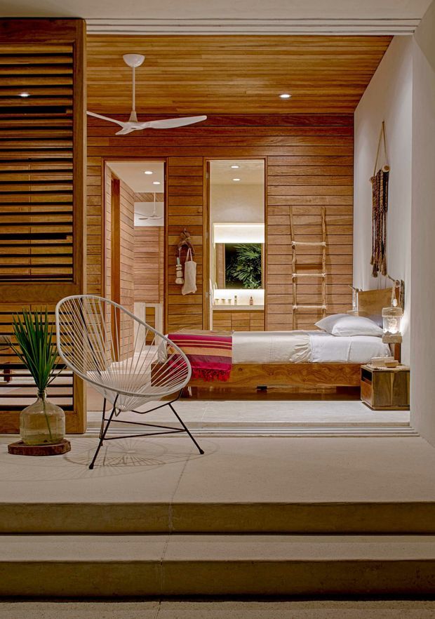 A warm, wood-lined bedroom viewed from a slightly lower stepped level. The room features extensive horizontal timber wall and ceiling panelling, a low built-in timber bed with white bedding and a colourful striped throw, mirrored wardrobe or dressing panels, a white ceiling fan, woven pendant and wall details, and a sculptural wire lounge chair. The palette is predominantly honey-toned wood, cream, white, and muted red accents, creating a relaxed tropical-modern atmosphere.