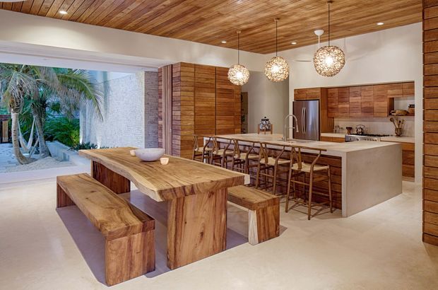 Open-plan kitchen and dining area with extensive warm timber detailing. A large live-edge timber dining table with matching benches sits in the foreground, while a timber kitchen island with bar stools occupies the rear. Timber cabinetry, wall panelling, and a timber-plank ceiling are paired with light neutral walls and flooring. Three spherical pendant lights hang above the island, and large openings connect the room to a lush outdoor courtyard or garden.