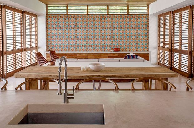 A bright open-plan kitchen and dining area with a large central island, integrated sink and gooseneck tap, timber dining table, curved timber chairs, and built-in bench seating. Warm timber-framed louvred windows surround the room, while a turquoise and terracotta patterned tile feature wall adds a strong decorative focal point. The space has a refined tropical-contemporary feel with abundant natural light.
