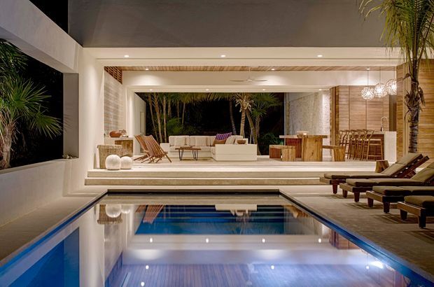 Night-time view of a refined indoor-outdoor pool courtyard. A rectangular swimming pool dominates the foreground, reflecting the illuminated white architectural pavilion, recessed ceiling lights, timber ceiling detailing and surrounding tropical planting. Beyond the pool are an outdoor lounge with armchairs and low tables, a dining area with timber furniture, decorative pendant lights, and large open glazed or framed openings connecting the spaces.
