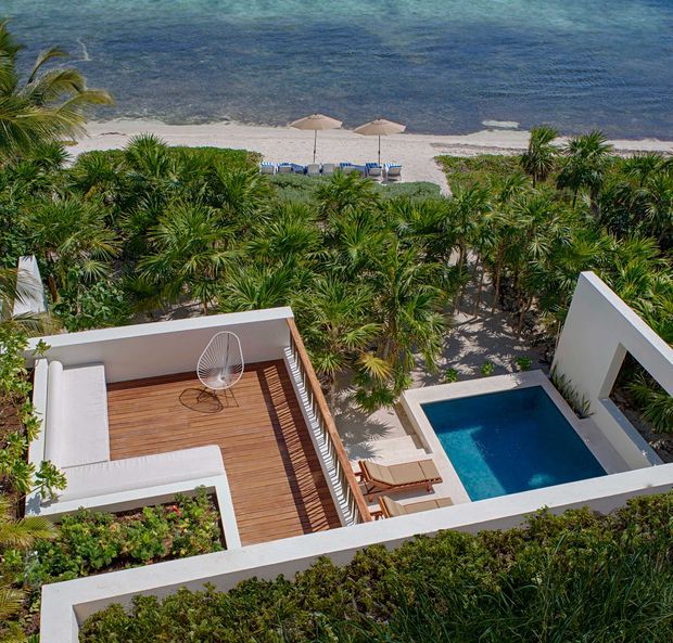 Elevated view of a tropical beachfront property with a white geometric terrace, timber deck, outdoor seating, private rectangular swimming pool, dense palm landscaping, and sandy beach with umbrellas and lounge chairs beyond. The scene has a clean contemporary resort-like design and strong indoor-outdoor connection.