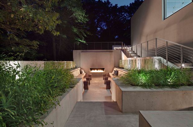 Night-time view of a contemporary landscaped courtyard and sunken outdoor lounge. A linear built-in fire feature anchors the space, with steps descending to a paved seating area surrounded by raised concrete planters and dense planting. The adjacent building has smooth rendered walls, large glazing, and simple horizontal metal balustrades.