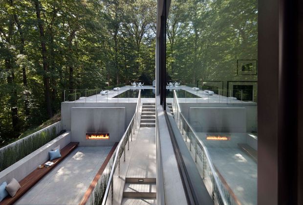 View from inside a modern home through a large glazed opening toward a split-level concrete terrace in a wooded setting. The terrace features a linear outdoor fireplace, built-in bench seating with cushions, glass balustrades, broad concrete surfaces, and a central stair descending between the levels. The glazing creates a strong reflection of the terrace and forest, emphasizing the indoor-outdoor connection.