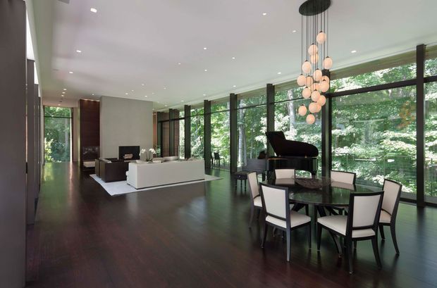 Spacious contemporary open-plan interior combining a living room and dining area. Dark timber flooring, white walls and ceiling, extensive black-framed floor-to-ceiling glazing, and a sculptural cluster pendant create a refined, light-filled setting overlooking mature greenery. A white sofa and low media unit define the living zone, while a dark dining table, upholstered chairs, and grand piano occupy the glazed side of the room.