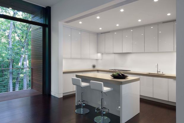A bright contemporary kitchen with glossy white full-height cabinetry, integrated appliances, a pale stone island and matching perimeter benchtops, and two white bar stools. Dark timber flooring contrasts with the white joinery, while a large floor-to-ceiling window brings in views of dense greenery. Recessed ceiling spotlights provide even illumination.