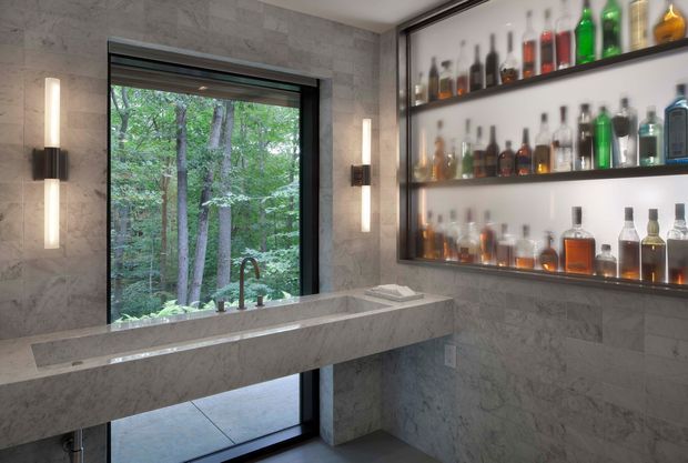 A sophisticated home bar with a long stone-clad counter and sink positioned beneath a large window overlooking trees. The walls and counter are finished in grey marble or stone, with dark-framed glazing, integrated open shelving displaying liquor bottles, and slim vertical wall sconces. The space has a refined, contemporary feel with strong natural views and layered lighting.