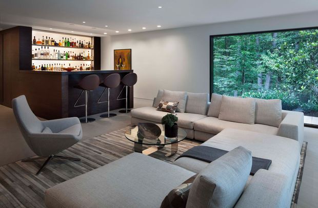 Contemporary entertainment lounge with a built-in dark timber home bar, illuminated bottle shelving, bar stools, and a large pale sectional sofa arranged around a glass coffee table. A wide black-framed window overlooks dense greenery, while recessed ceiling lighting and grey-toned timber flooring create a refined, comfortable interior.