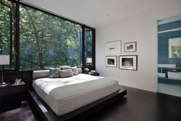 Contemporary bedroom with a low-profile dark timber platform bed, crisp white bedding, and patterned accent cushions. Full-height corner glazing with dark frames opens the room visually to dense greenery, while a minimalist gallery wall and dark flooring add contrast. An adjoining blue-tiled bathroom is visible through the open doorway.
