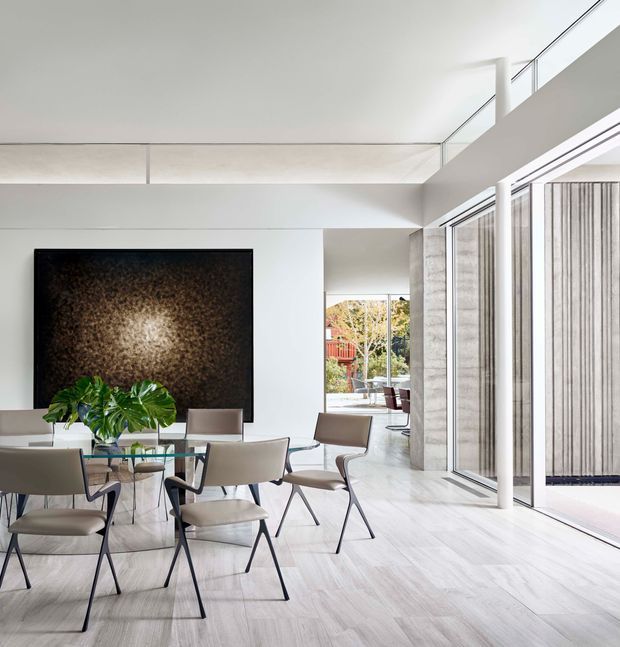 A bright, open-plan dining area with a glass dining table and several modern upholstered chairs. The room has pale timber-look flooring, white walls and ceiling planes, large floor-to-ceiling glazed doors, and a dramatic dark textured feature artwork or wall panel. A planted indoor arrangement sits on the table, while a glimpse of an outdoor terrace and garden is visible beyond the glazing. A high-level opening and glass balustrade suggest a double-height or mezzanine-connected space.