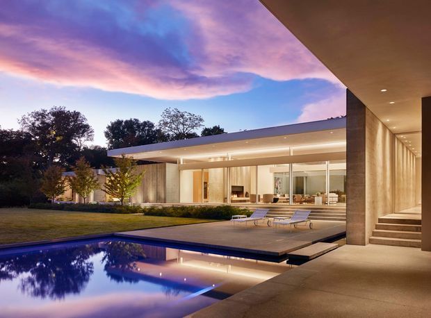 Twilight view of a luxurious modern single-storey residence with broad floor-to-ceiling glazing, a projecting flat roof, warm recessed lighting, landscaped lawn, and a reflective rectangular swimming pool. A covered terrace and stepped entry frame the right side of the composition.