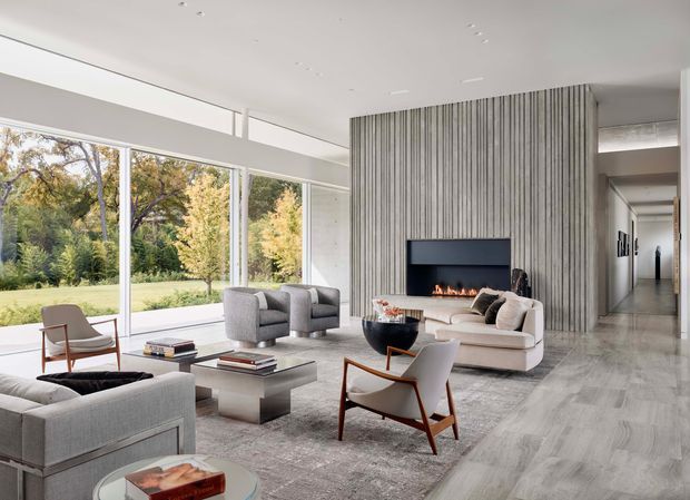 A spacious, refined contemporary living room with full-height glazing overlooking landscaped grounds. The room features a large grey sectional sofa, sculptural lounge chairs, a low coffee table, a linear fireplace set into a dramatic vertically ribbed feature wall, polished pale flooring, and a long corridor extending beyond the main space.