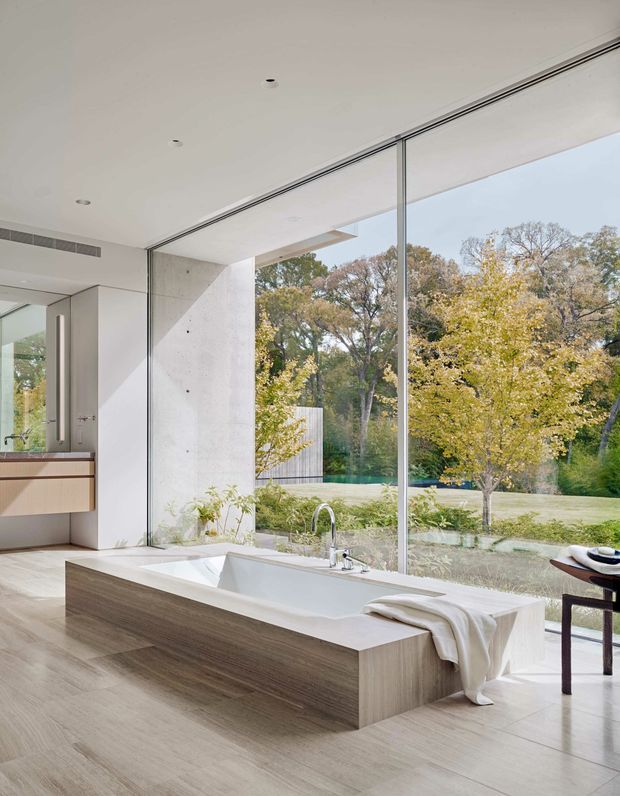 Bright contemporary bathroom with a freestanding-style rectangular bathtub integrated into a timber-look surround, set beside full-height glazing overlooking a landscaped garden and pool. The room features pale stone or concrete wall surfaces, light wood-look flooring and cabinetry, minimalist tapware, recessed ceiling lights, and a large frameless-looking window wall.
