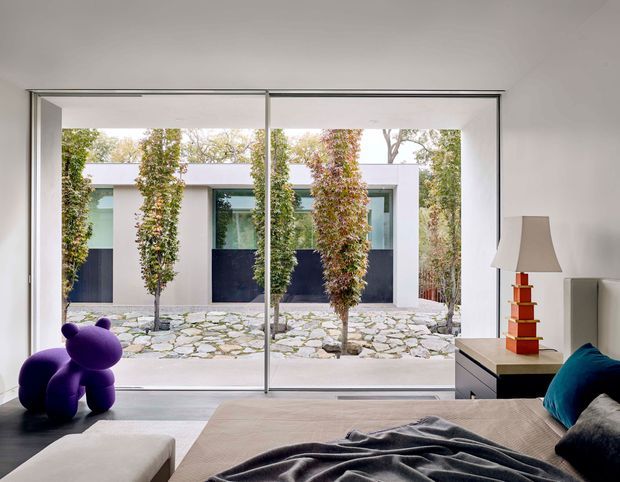 A contemporary bedroom viewed toward a private courtyard through full-height sliding glass doors. The room has a neutral upholstered bed, pale walls, a dark bedside cabinet, a sculptural purple chair, and a colourful stacked-base table lamp. Outside, a stone-paved courtyard with slender planted trees and a modern low white building is visible.