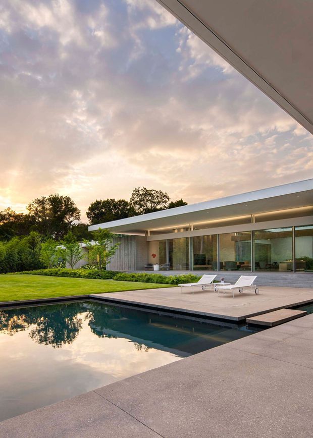 Exterior view of a low-slung modern residence opening onto a large paved terrace and reflecting swimming pool. The home features a broad flat roof with deep overhangs, extensive floor-to-ceiling glazing, and a restrained grey and natural timber material palette. Manicured lawn and mature planting frame the pool, while the sunset sky reflects in the dark water.