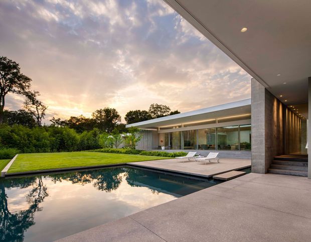 A contemporary modern residence viewed from a covered outdoor area, overlooking a rectangular swimming pool, paved terrace, manicured lawn, and landscaped garden. The low-slung house has extensive floor-to-ceiling glazing, a flat roof, concrete or stone-clad structural walls, and a recessed illuminated entry stair. The scene is photographed at sunset, with the pool reflecting the sky and surrounding trees.
