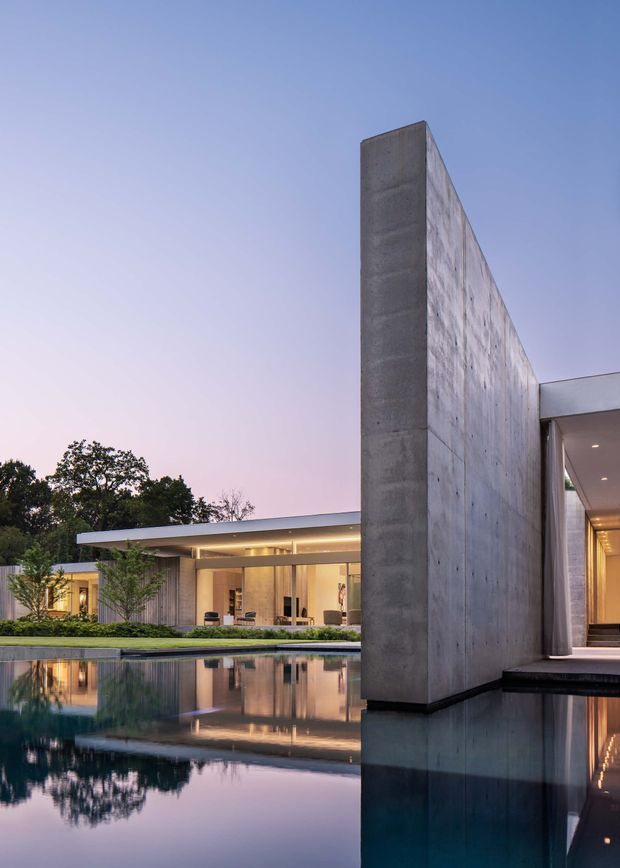 Dusk exterior view of a refined modern residence with strong horizontal roof planes, extensive glazing, and a tall exposed concrete feature wall. The house is arranged around a reflective swimming pool, with warm interior lighting visible through the glass and landscaped planting along the pool edge.