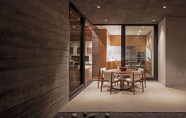 A refined contemporary dining area with a timber dining table and four chairs, opening directly onto a warm timber-lined kitchen. Full-height black-framed glazing and sliding doors connect the spaces, while a dark textured concrete wall and concrete ceiling add an understated architectural character. Recessed lighting and warm timber cabinetry create a calm, sophisticated atmosphere.
