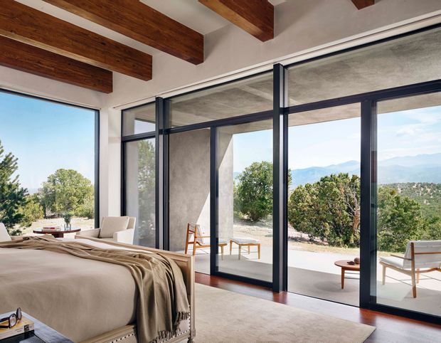 A bright, refined bedroom with a large upholstered bed in the foreground, warm timber ceiling beams, and expansive dark-framed floor-to-ceiling glazing opening onto a terrace and mountain landscape. The room uses a restrained palette of cream, beige, white, dark timber, and charcoal window frames, with minimalist seating and a calm indoor-outdoor connection.