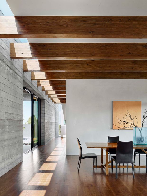 A contemporary dining area in a long, open-plan interior. Dark timber ceiling beams run across the room, creating a strong linear rhythm over a polished dark timber floor. The space combines a raw horizontal concrete wall with smooth white plaster walls and full-height glazing along the left side. A timber dining table with black dining chairs sits beneath minimalist artwork and a large glass vessel, while sunlight creates rectangular patches across the floor.
