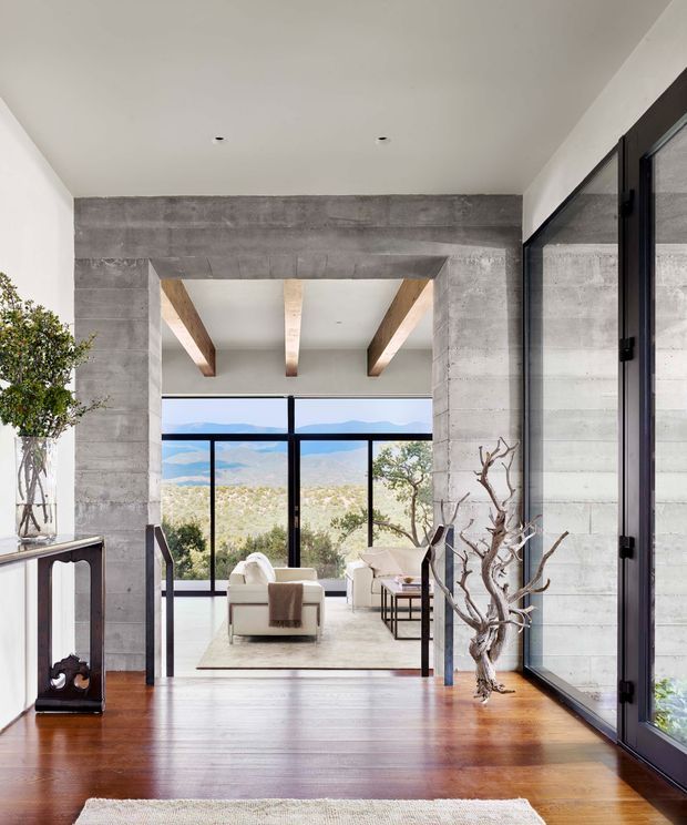 A bright, modern interior view looking through a concrete-framed opening into a living room. Exposed concrete walls and beam, timber ceiling beams, dark-framed floor-to-ceiling glazing, polished warm timber flooring, and a distant mountain landscape create a strong indoor-outdoor connection. A small console, lounge seating, side tables, and sculptural branch decor are visible.