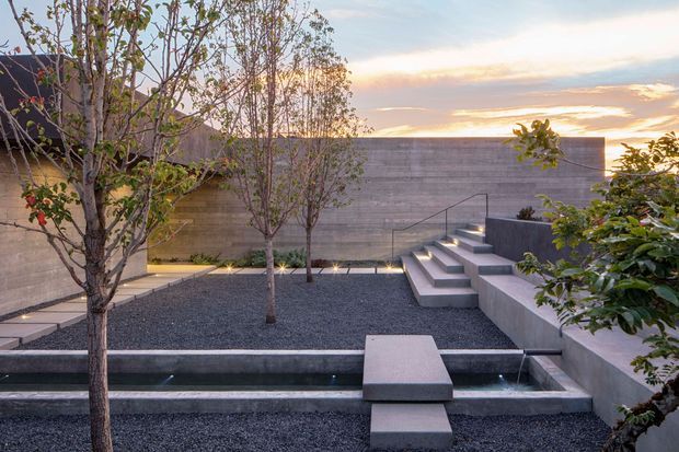 A minimalist landscaped courtyard at sunset, featuring a long narrow reflecting pool with concrete stepping blocks, dark gravel ground cover, sculptural concrete stairs and raised seating or planter edges, mature trees, and a tall horizontal rendered concrete boundary wall. Recessed lighting accents the paths and water feature.